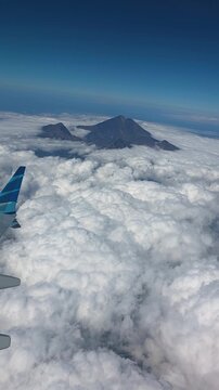 Mount Rinjani rises through a blanket of white clouds beside an aircraft wing during a flight, aerial backdrop, vertical