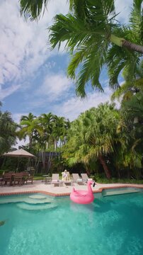 Vertical Screen: Woman relaxing by the pool on a sun lounger in a straw hat, low angle under a leaning palm. Pink flamingo float on turquoise water, patio table, umbrella and tall palms on a sunny day