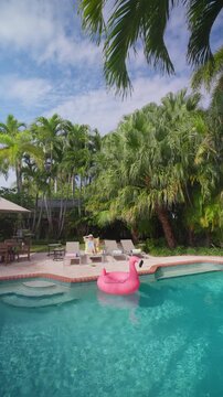 Vertical Screen: Woman relaxing by the pool on a yellow towel holds a drink and her straw hat. Pink flamingo float on turquoise water, tall palms, grill, patio table and gray loungers on a sunny day.