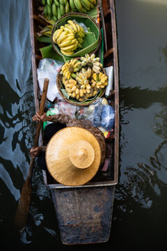 Traditional floating market in Bangkok, Thailand, woman local vendor in straw hat sells fresh bananas from boat, exotic boat shop offering and selling fresh tropical fruits from wooden boats in Asia.