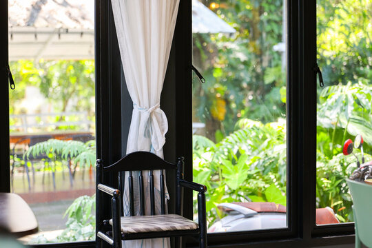 Black wooden chair beside open window with white curtain, tropical garden, natural light, and quiet home interior creating calm reading space