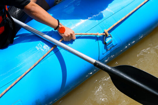 Blue inflatable raft with paddler gripping rope and oar above muddy river water, outdoor adventure scene under bright sunlight and strong shadow contrast