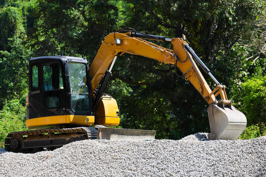 Yellow excavator with metal bucket resting on gravel mound beside dense green forest, showing construction equipment ready for outdoor earthwork and material handling