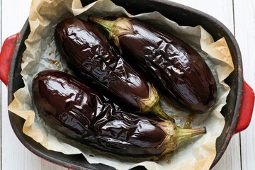 Naklejka na ściany i meble Three whole purple eggplants bake in metal pan with parchment paper. Roasted aubergines rest on baking sheet for healthy meal preparation. Fresh organic vegetables cook in oven for delicious