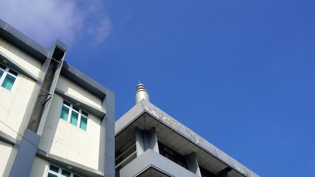 Cream and gray building with green glass windows stands beside lower rooftop with metal tower, under saturated blue sky with faint wispy cloud at upper left.