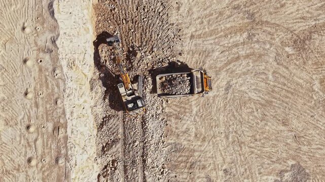 A loader and haul truck move rock across a quarry work area during the day, with tracks and cut earth visible across the site.