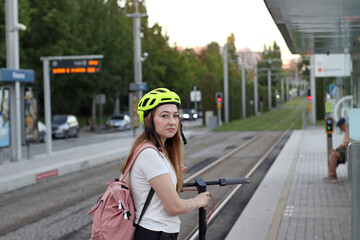 Young woman in a yellow helmet with a pink backpack and an electric scooter on a metro platform near rails, waiting for a train. Suitable for urban mobility, transport, travel and lifestyle themes. © Evgeniia