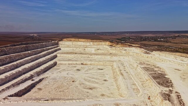 A quarry with stepped excavation levels and pale exposed ground extends across open terrain, with distant settlements visible along the horizon in daylight.