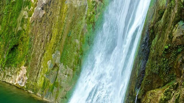 Cascading water falls dramatically over moss-covered rock cliffs into a clear turquoise river pool during daytime sunshine.