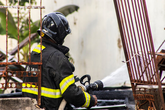 A firefighter stands amidst rubble and thick smoke, directing a high-pressure water hose to extinguish a major structural fire destroying a house in Marilia, Sao Paulo state