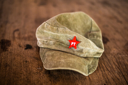A close-up shot displays a traditional red star pin featuring the acronym "PT" of the Brazilian Workers' Party pinned to an olive green corduroy military-style fatigue cap, resting