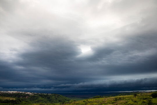 Dark, ominous storm clouds and tempest weather sweeping across a lush green valley and rural landscape with a small town in the countryside of Brazil.