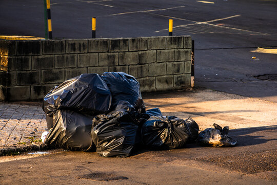 Black plastic trash bags piled up on a public street sidewalk waiting for garbage collection in the urban area of Brazil. Concept of waste management, environmental pollution, 
