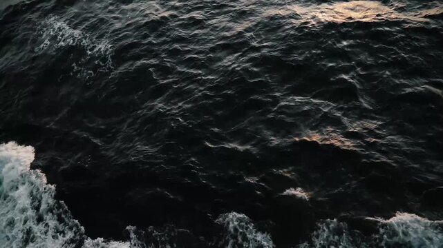 Aerial drone view of dark ocean water with white foamy waves crashing under golden-hour light