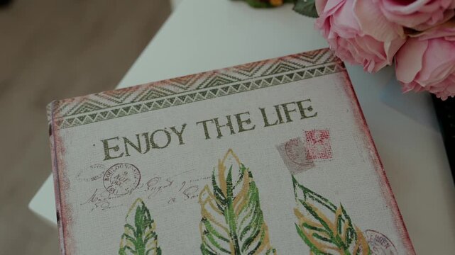 Book shaped storage box printed with Enjoy the Life, feathers and postmarks, lying on a table beside pink roses.