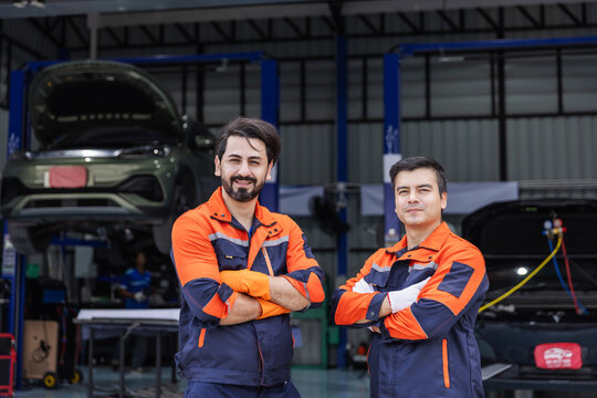 Two confident EV mechanics wearing orange and navy uniforms stand with arms crossed and look at the camera in an automotive workshop.