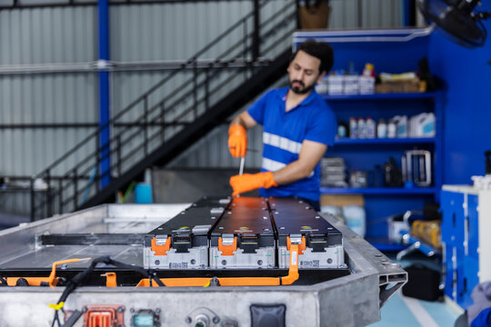Technician wearing a blue work shirt and orange gloves uses a hand tool to tighten fasteners during electric vehicle battery module assembly.