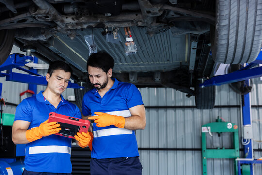Two automotive technicians wearing blue work shirts and orange gloves review a diagnostic tablet beneath an electric vehicle raised on a service lift. 
