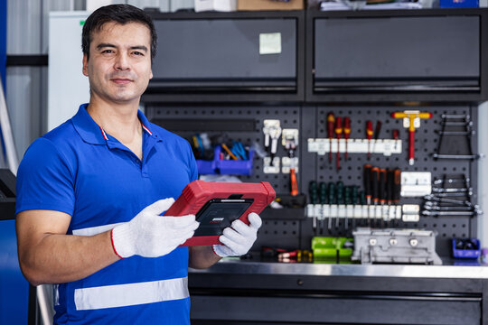 Automotive technician wearing a blue work shirt and protective gloves holds a red diagnostic tablet while looking at the camera.