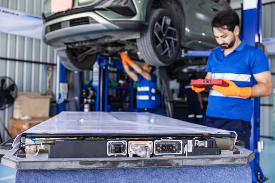 Technicians inspect an electric vehicle after flood exposure in an automotive workshop. A removed battery pack sits in the foreground while one technician holds a diagnostic tablet.