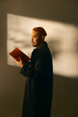 Profile portrait of a thoughtful bearded man in a dark coat and orange beanie reading an open book, illuminated by warm indoor sunlight and soft shadows, expressing study, education, and calmness. © Francis