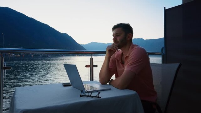 Focused remote worker using laptop on tranquil lakeside balcony at dusk, enjoying peaceful outdoor workspace with scenic mountains and calm water in background