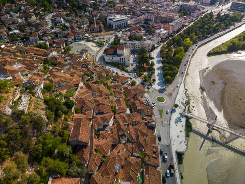 Berat Albania spreads across riverfront in broad aerial perspective, with terracotta roofs packed around town streets. Riverside road curves past trees, plaza and shallow stony waterway.