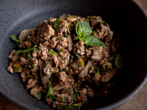 Thai spicy minced pork larb salad topped with fresh basil leaves, chili and herbs, served in a black bowl on a rustic wooden table, top view of northern Thai food