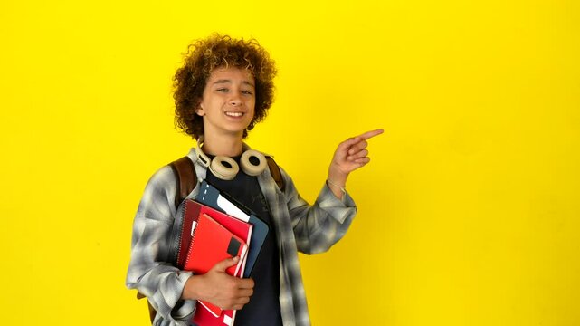 Cheerful teenage student with a backpack and notebooks smiling and pointing his finger at a copy space on a vibrant yellow background, presenting information for an educational concept