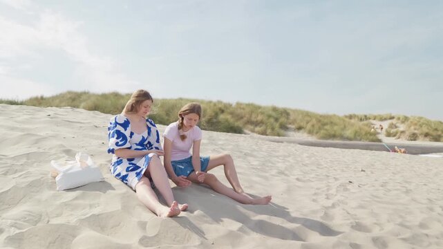 A woman and girl are sitting on the sand and talking slow motion sandy dunes by the sea, 35 years old woman and 15 years old girl wide shot