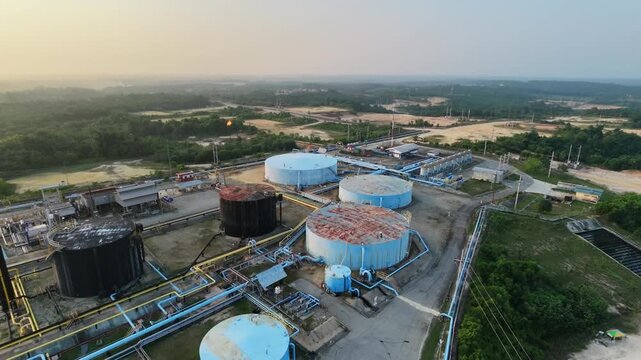 Aerial overview of an energy production site featuring cylindrical storage tanks and waste management ponds under soft daylight. Petroleum industry, fuel distribution, and infrastructure.