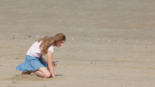 Girl digging sand and holding a smartphone slow motion by the sea