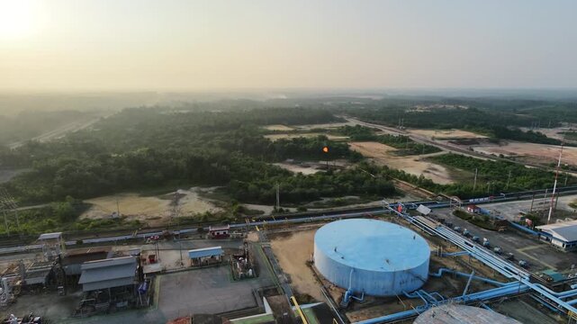 Aerial overview tracking across a sprawling oil and gas refinery plant under a hazy horizon. Highlights large industrial tanks, processing facilities, and complex technical piping.