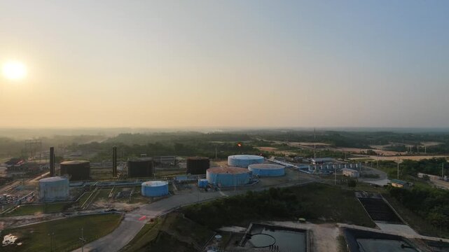 Aerial overview tracking across a sprawling oil and gas refinery plant under a hazy horizon. Highlights large industrial tanks, processing facilities, and complex technical piping.