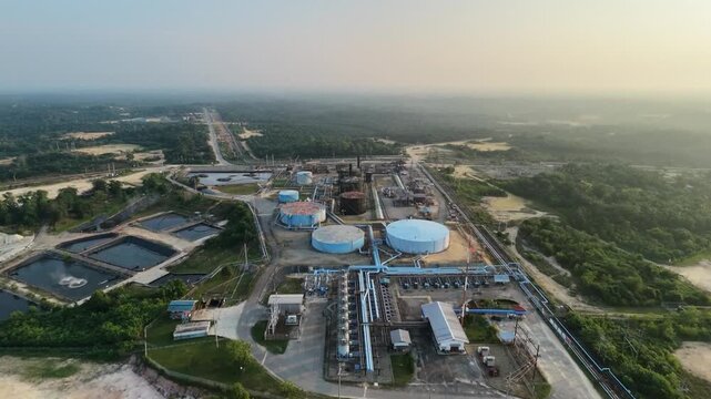 Panoramic aerial view of a sprawling petroleum facility and energy plant during twilight. Includes distinct blue and black storage silos, processing units, and a series of industrial wastewater lagoon