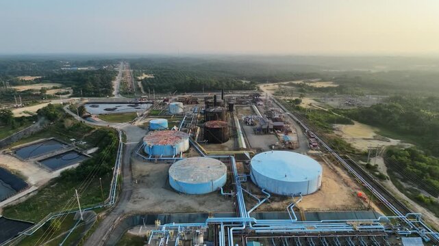 Panoramic aerial view of a sprawling petroleum facility and energy plant during twilight. Includes distinct blue and black storage silos, processing units, and a series of industrial wastewater lagoon