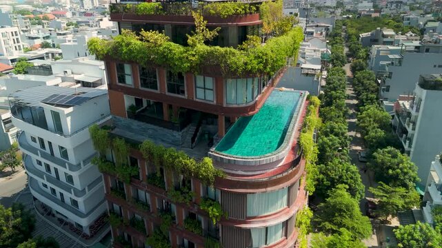 Green rooftop terraces and pool crown modern urban tower, offering shade among dense city buildings. Aerial view highlights cooling vegetation and reflective water as heat conscious design. Danang