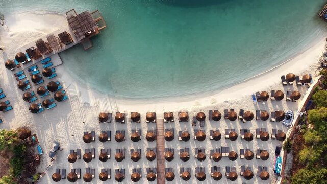 Aerial view of Ksamil beach featuring rows of sun loungers and straw umbrellas on white sand along the turquoise Ionian Sea in Ksamil, Vlore County, Albania.
