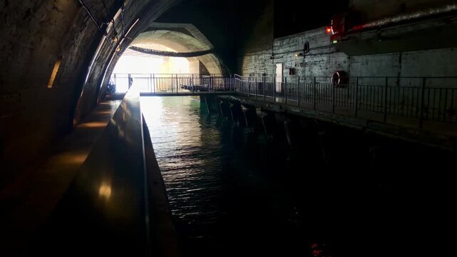 A dimly lit, atmospheric interior view of the underground canal. The space features a massive arched concrete ceiling and walkways along the sides. The scene is illuminated by rows of warm, yellowish 