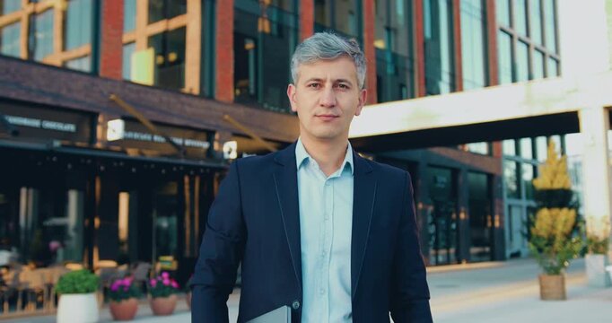 A confident, successful bearded businessman in formal suit stands outdoors with arms crossed, smiling and looking directly at the camera against the backdrop of a modern glass office building.