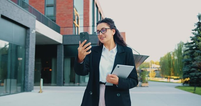 Young professional woman walking outside modern office building checking smartphone and holding laptop, smiling and speaking during daytime urban commute, three-quarter portrait view