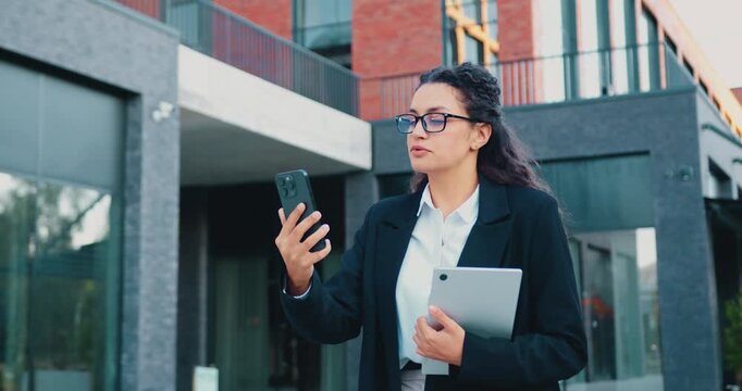 Cheerful Caucasian woman holding smartphone and recording vlog while walking outdoors in city, smiling and talking during video call. Influencer lifestyle concept.