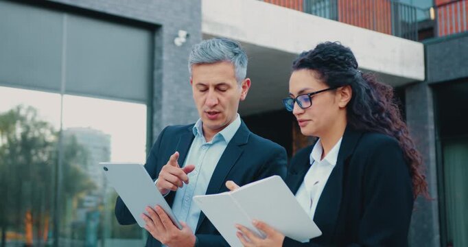 Close-up of a businessman with a laptop and an elegant young businesswoman using a smartphone outside a modern office building, discussing ideas and analyzing data together. Slow motion
