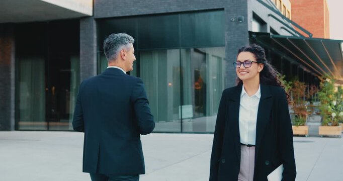 Two colleagues of adult man and young woman, walk toward each other outside a modern office building, smiling cheerfully, exchanging a high five, then parting ways to continue their day