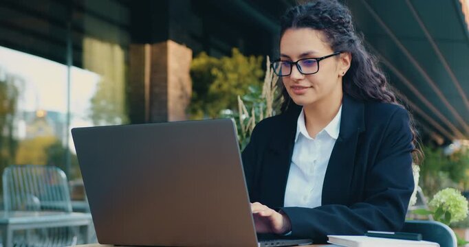 Smiling businesswoman working on a laptop outdoors in an urban setting. Concept of finance, corporate management, digital banking, fintech solutions, financial analysis, and modern business