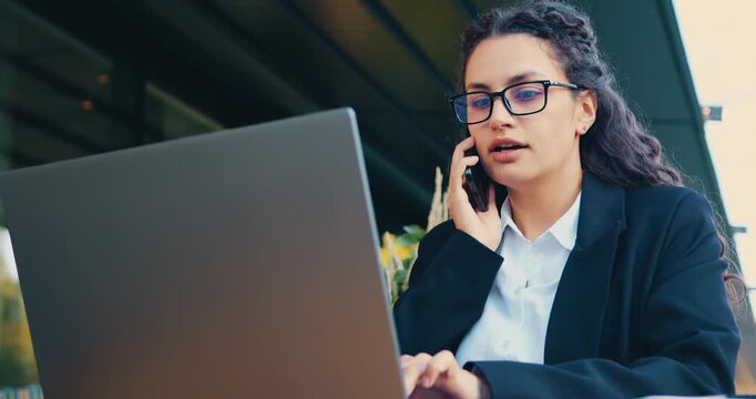 Busy businesswoman sitting on an outdoor terrace during lunch break, talking on smartphone, drinking coffee, and typing on laptop. Professional manager focused on remote work, multitasking, and modern