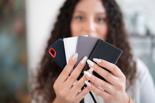 A young woman with curly hair holds up a fan of several portable external solid state drives in her hands, showing the data storage devices to the camera indoors.