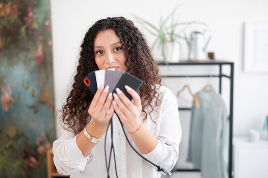 A young woman with curly brown hair holds up several colorful smartphone cases in a bright modern room. She smiles softly at the camera while showing the accessories.