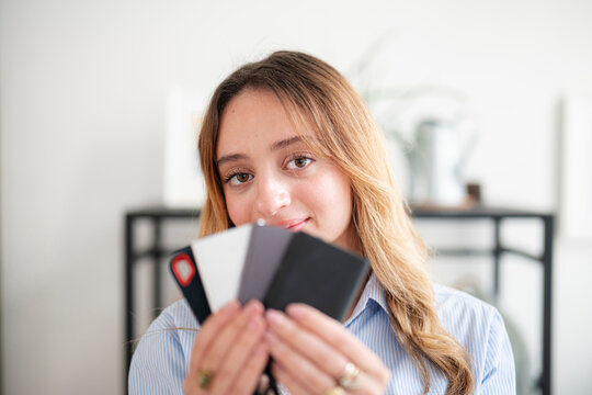 A young blonde woman wearing a blue striped shirt smiles gently as she holds up a fan of small colorful cardholders in her hands inside a bright indoor workspace.