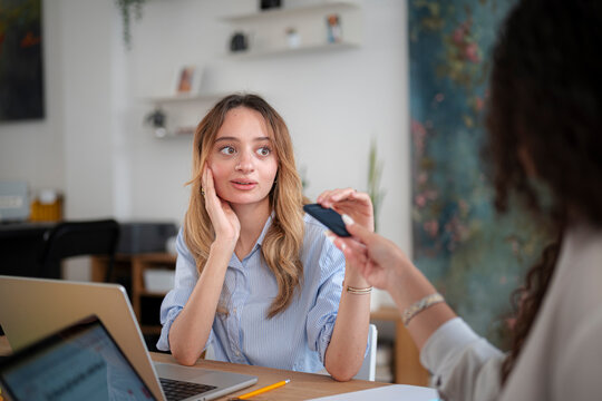 Two female colleagues collaborate in a bright office. One blonde woman sits at a desk with laptops and looks shocked while another coworker hands her a smartphone.